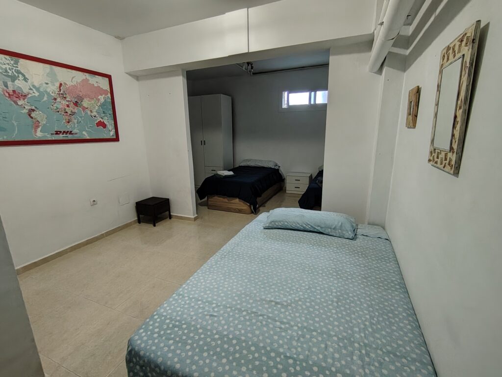 La habitación cuenta con una cama individual con colchón y almohada, y una pequeña mesa de noche. La pared de fondo es