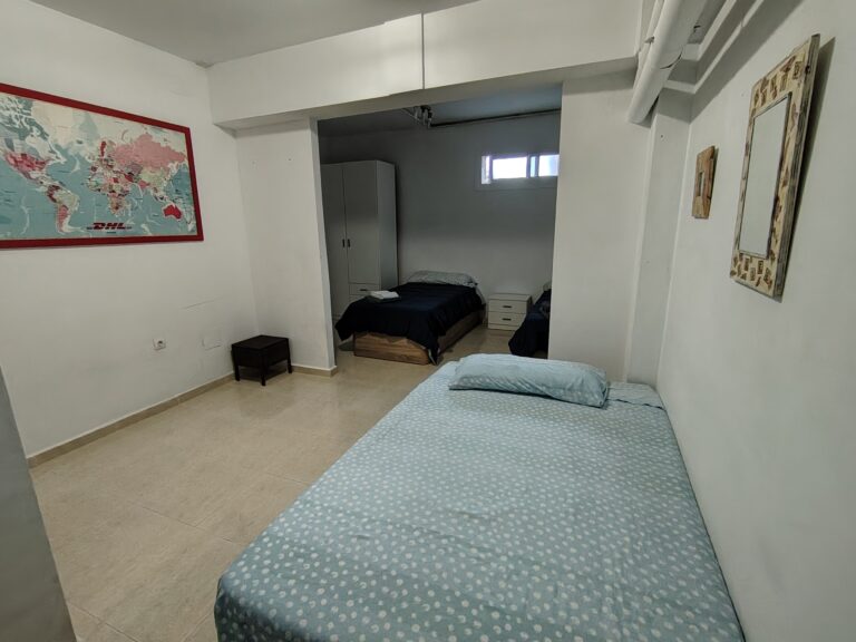 La habitación cuenta con una cama individual con colchón y almohada, y una pequeña mesa de noche. La pared de fondo es