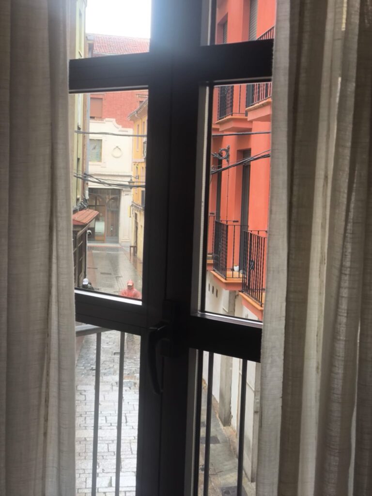 Una ventana con cortinas abiertas que da a un callejón estrecho y empedrado de una ciudad, probablemente León