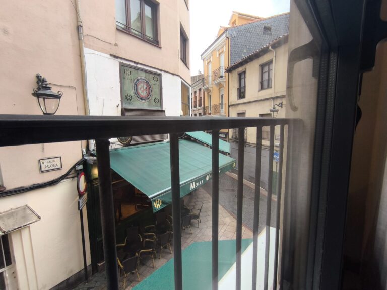 Una vista desde una ventana de un balcón, con una calle estrecha y un restaurante en el fondo. El restaurante tiene