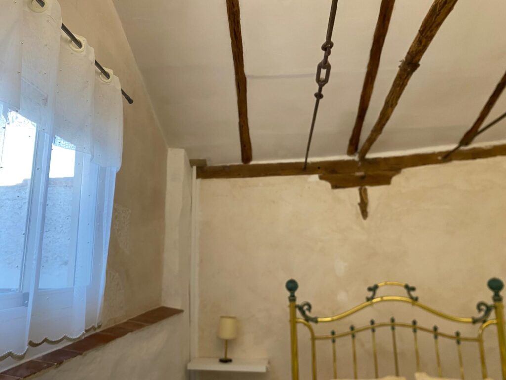 Una habitación con techo de madera y vigas visibles, paredes de color blanco y un colchón de metal con una estructura