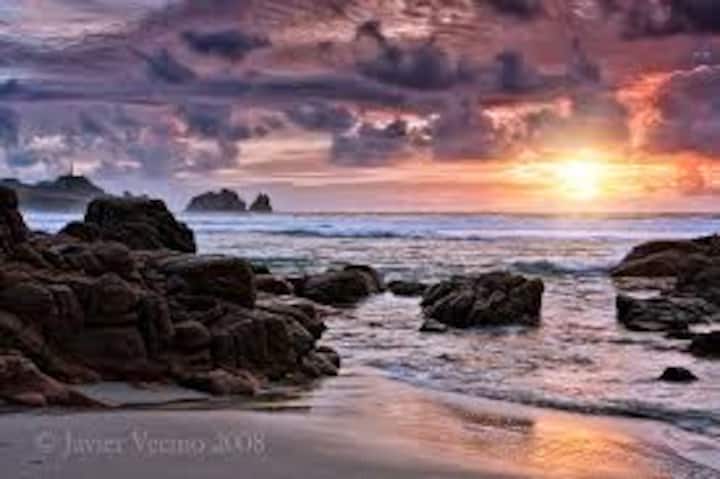 Una playa con rocas en la orilla, con un cielo nublado y un sol que se asoma entre las nubes