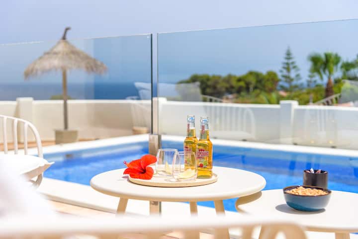 Una terraza con una mesa redonda y dos botellas de cerveza, dos vasos de vino blanco y un hibisco rojo