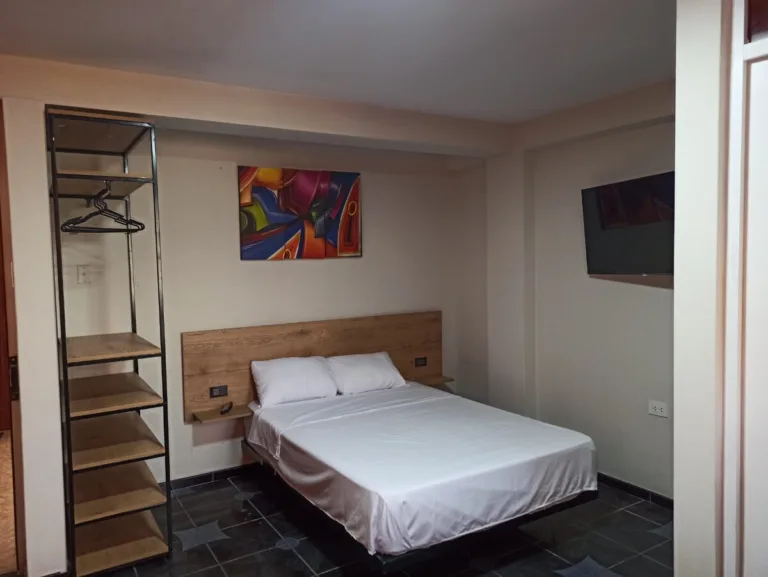 Una habitación con colores neutros, como el beis y el blanco, un televisor en la pared y un colgador para ropa